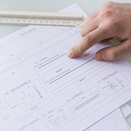 Primo piano della mano di una persona che indica un punto specifico su un documento tecnico ricco di disegni industriali e schemi quotati.